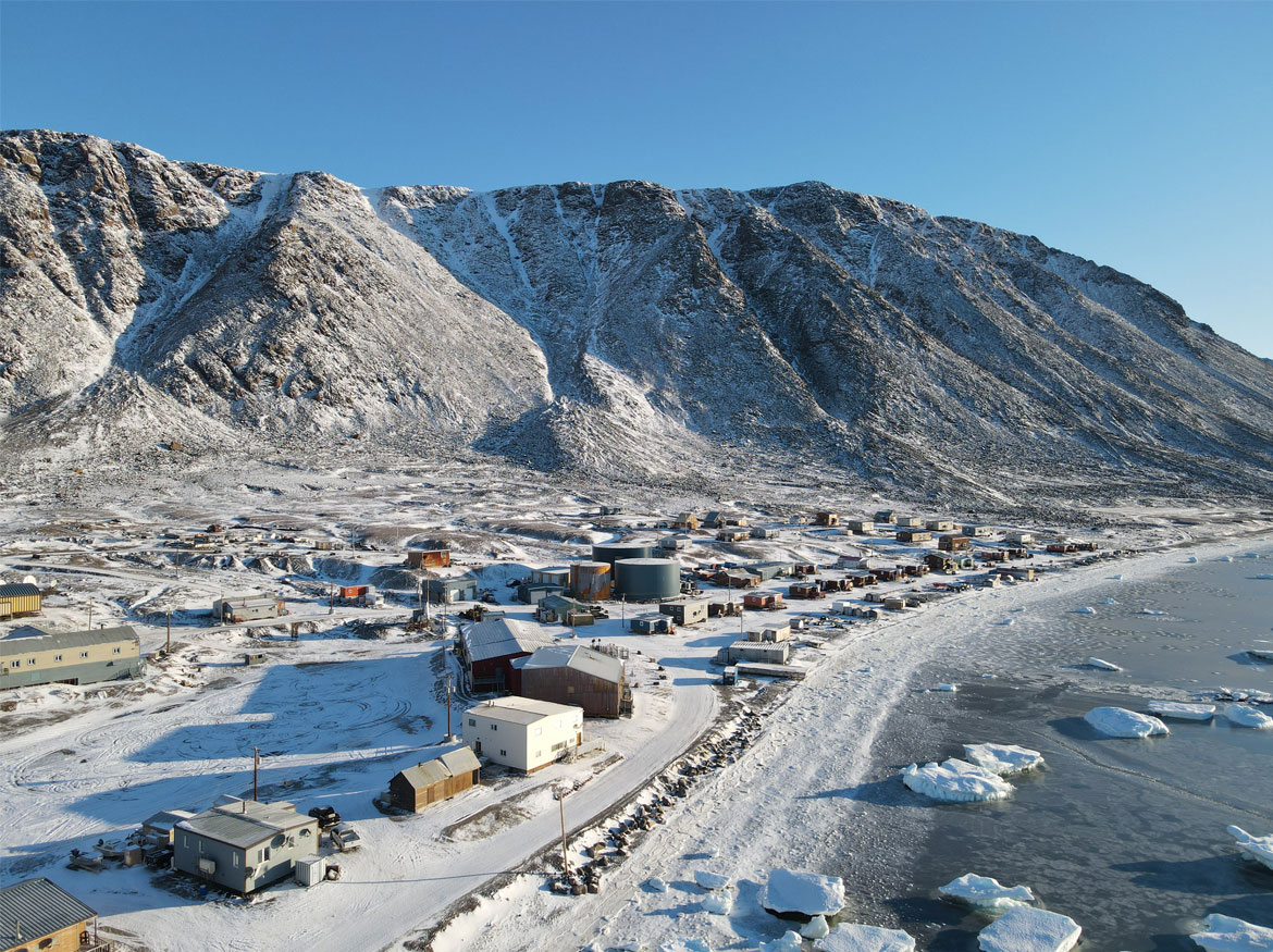 Vue aérienne de la communauté de Grise Fiord, au Nunavut, avec les montagnes à l’arrière, les bâtiments au centre et les eaux côtières glacées à l’avant.