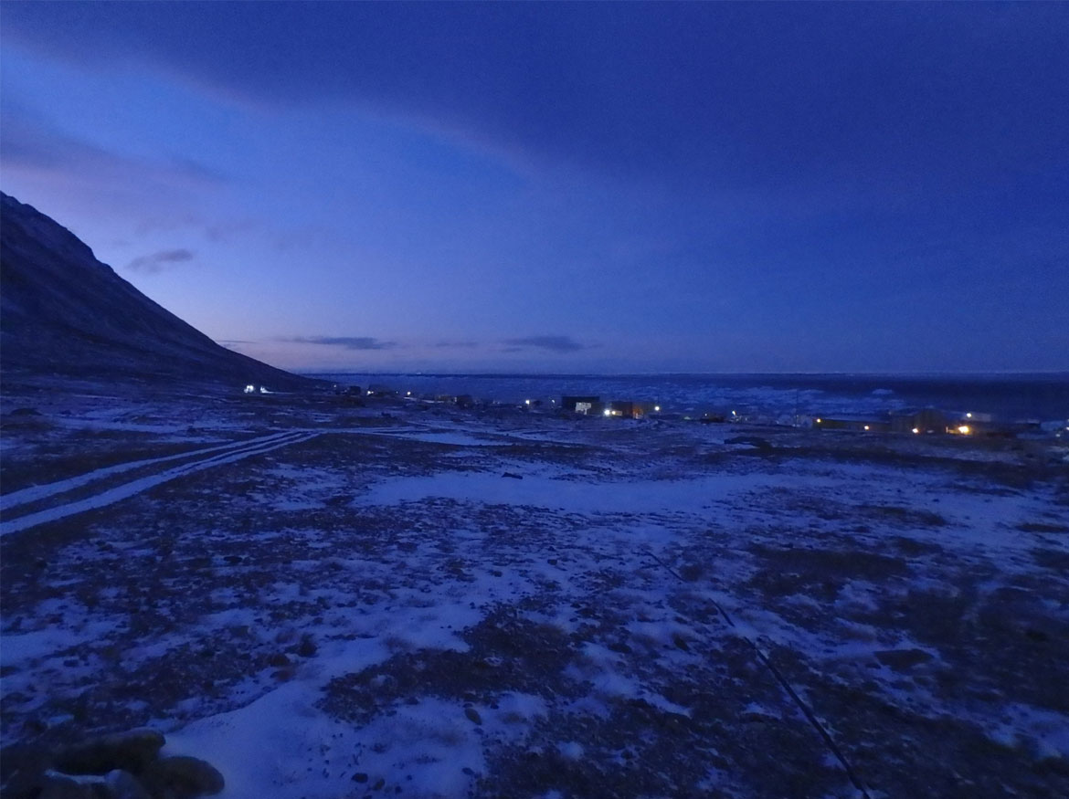 To the left, the sun is rising from behind a mountain and the lit hamlet of Grise Fiord, Nunavut is in the distance.