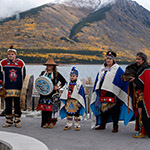 Cinq (5) danseurs portant des costumes traditionnels se tiennent devant un plan d'eau derrière lequel se trouvent des montagnes aux pics enneigés.